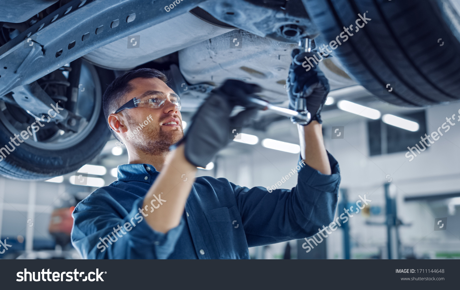 Portrait Shot of a Handsome Mechanic Working on a Vehicle in a Car Service. Professional Repairman is Wearing Gloves and Using a Ratchet Underneath the Car. Modern Clean Workshop. #1711144648