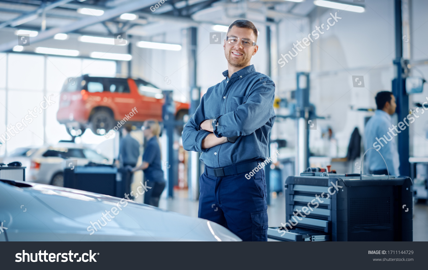 Handsome Car Mechanic is Posing in a Car Service. He Wears a Jeans Shirt and Safety Glasses. His Arms are Crossed. Specialist Looks at a Camera and Smiles. #1711144729