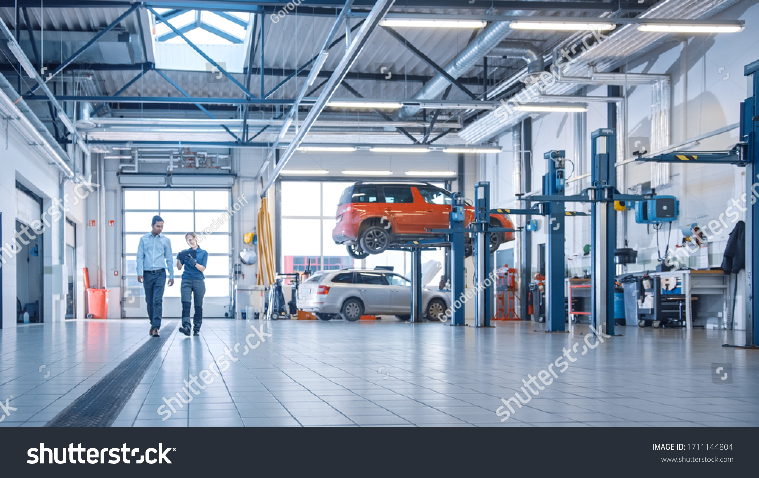 Female Mechanic Checks Diagnostics Results on a Tablet Computer and Explains a Vehicle Breakdown to a Manager. Car Service Employees Talk while Walking in a Garage. Modern Clean Workshop. #1711144804