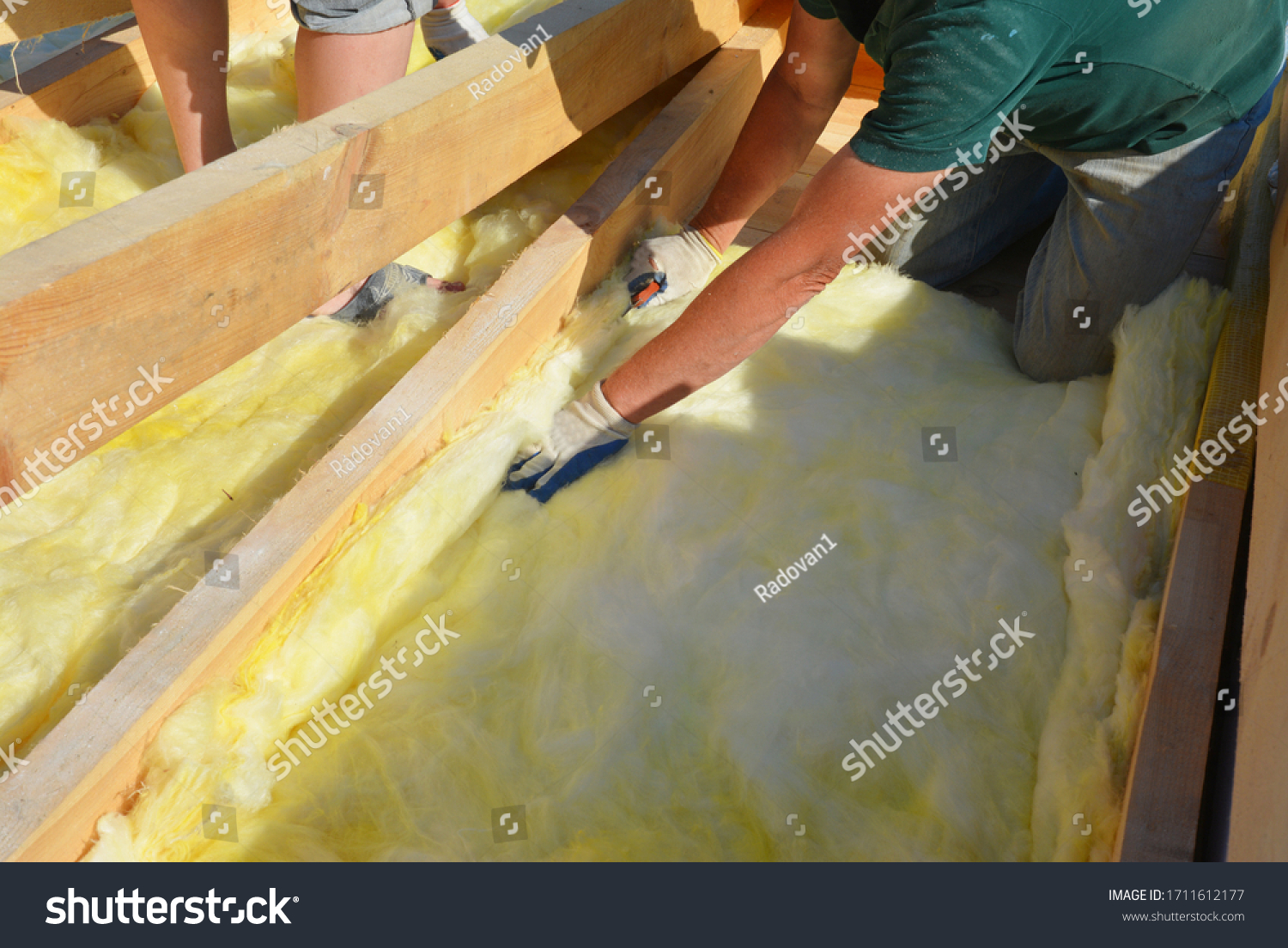 Roofing construction: building contractors in protective gloves are installing glass wool sheets, batts on the rooftop below trusses for thermal insulation of the rooftop. #1711612177