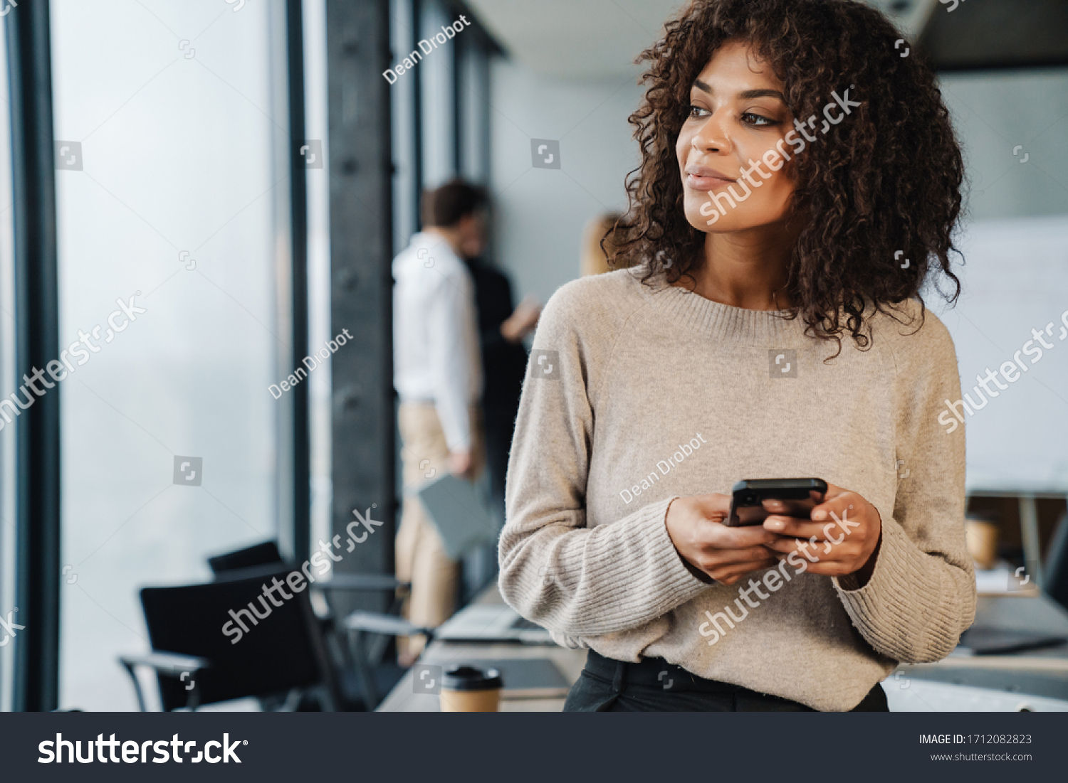 Beautiful african smiling young smart businesswoman standing in the office with group of colleagues on the background, using mobile phone #1712082823