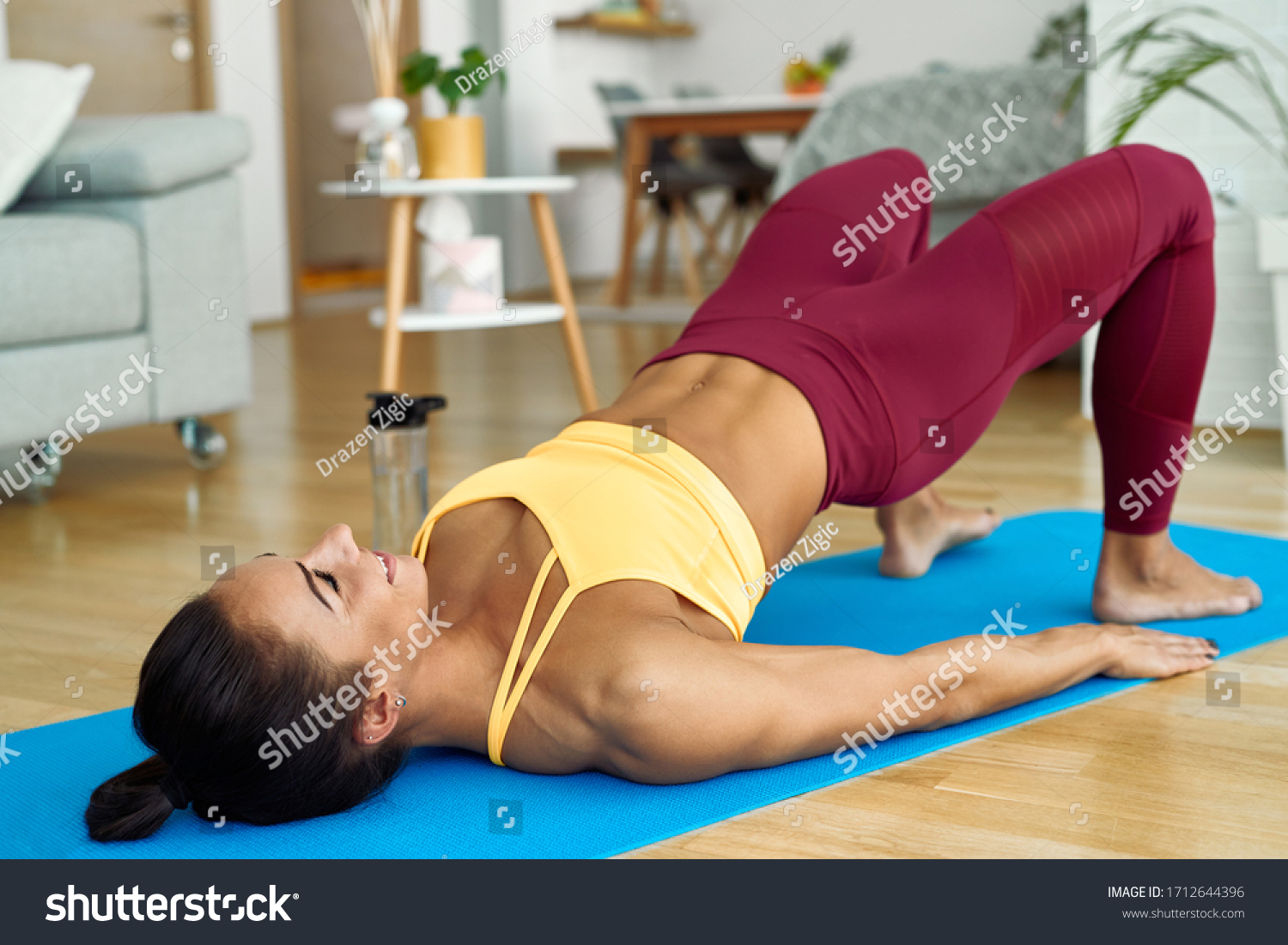 Young athletic woman lifting her hips while doing glute bridge exercise on the floor at home.  #1712644396