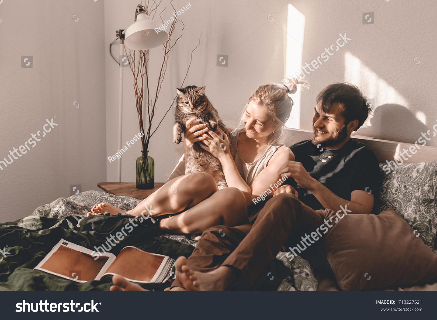 Happy couple is spending time in bed. Weekend leisure time. Time together. Stay home. Relax in bed. Modern bedroom. Read, eat, laugh. Lying in bed together. Bright modern bedroom. Kitty cuddles. #1713227521