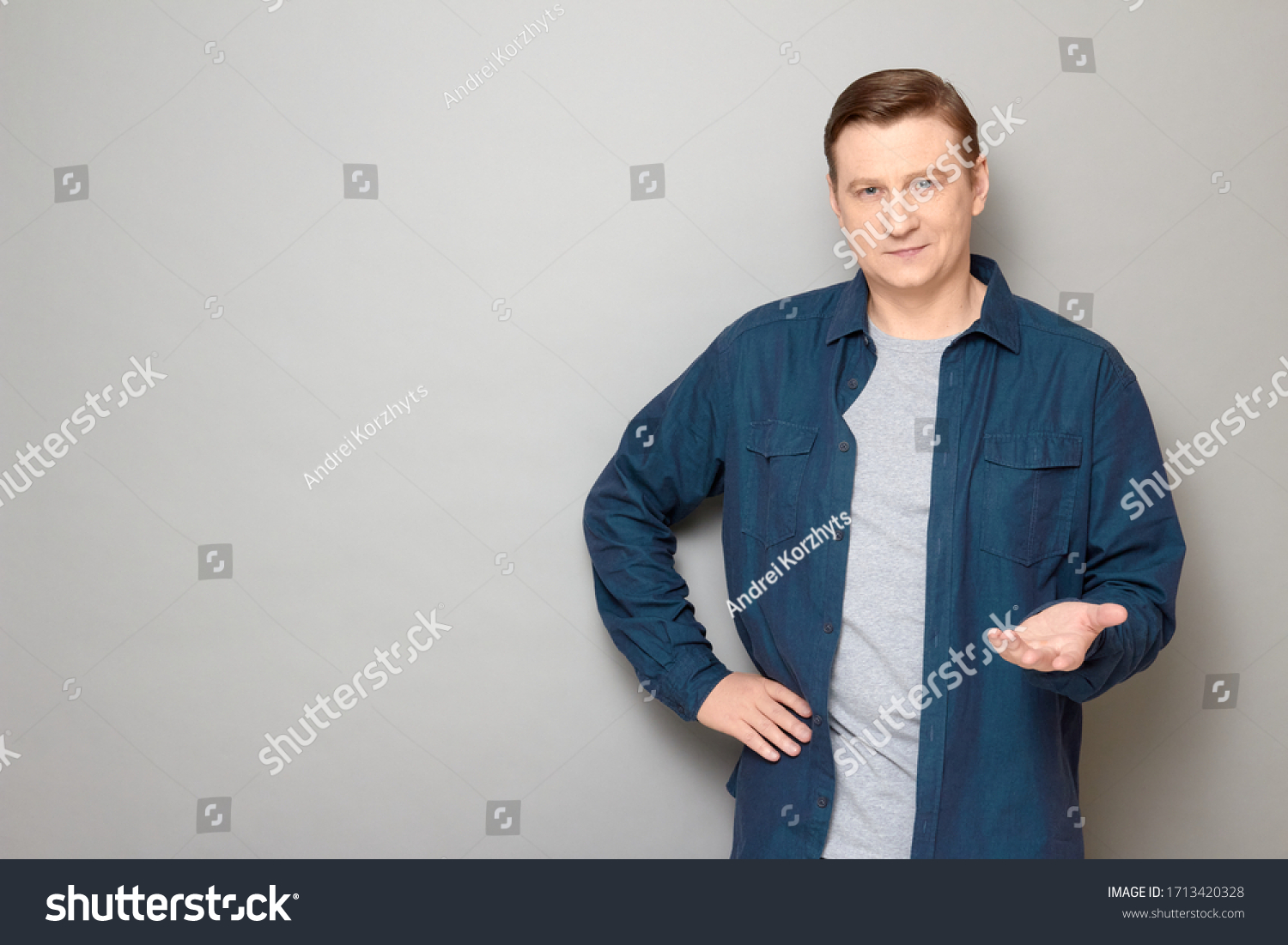 Studio portrait of friendly man wearing blue shirt, smiling, stretching one hand like offering product, asking your opinion or waiting your decision, standing over gray background, copy space on left #1713420328