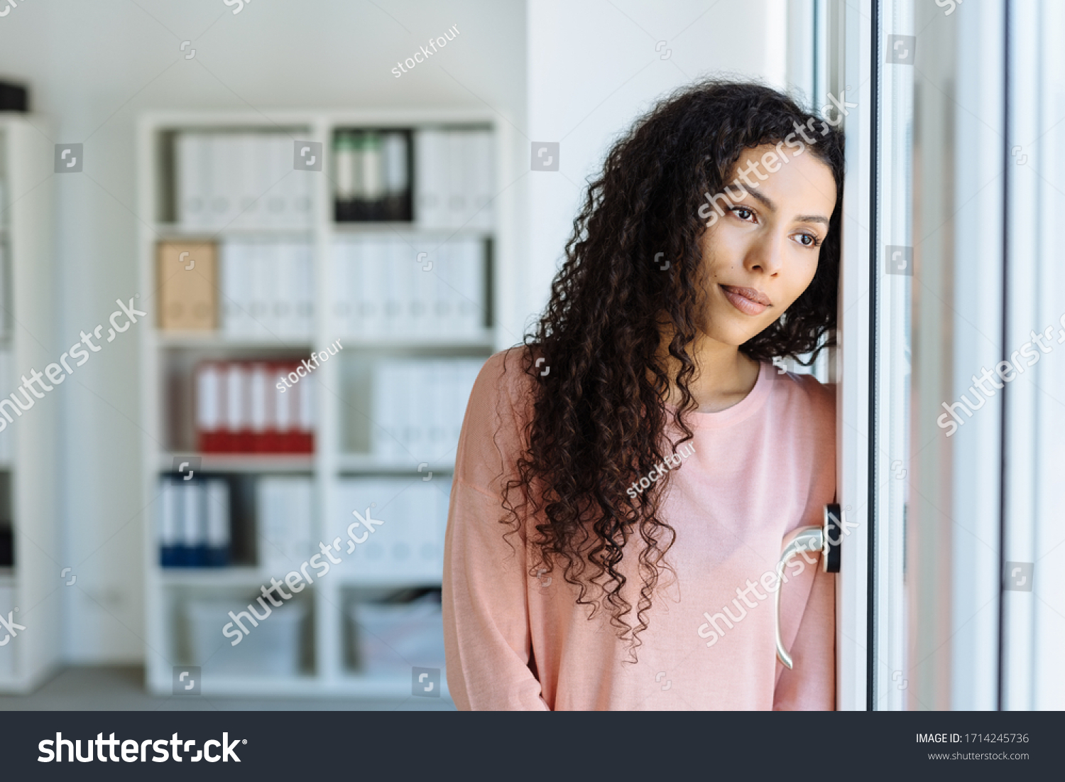 Young woman staring longingly through a window with a sad faraway expression as she leans against a wall in an office #1714245736
