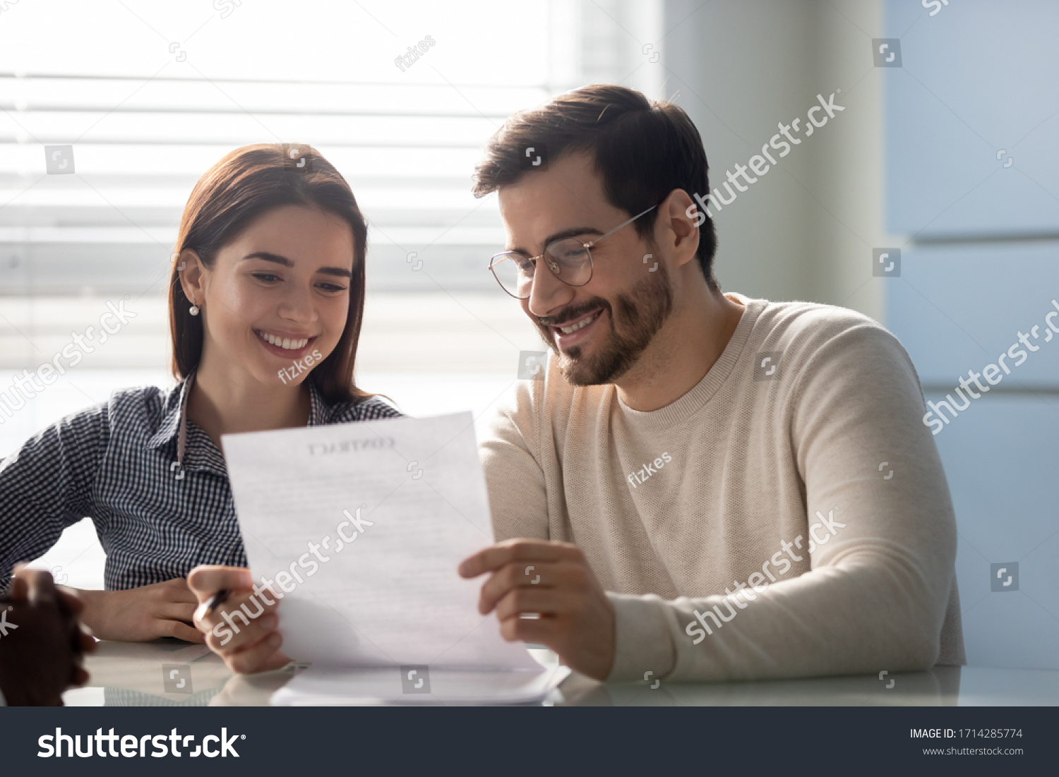 Young successful couple, man and woman reading business document at mortgage negotiating. Happy smiling husband in glasses and wife making deal together in broker office. #1714285774