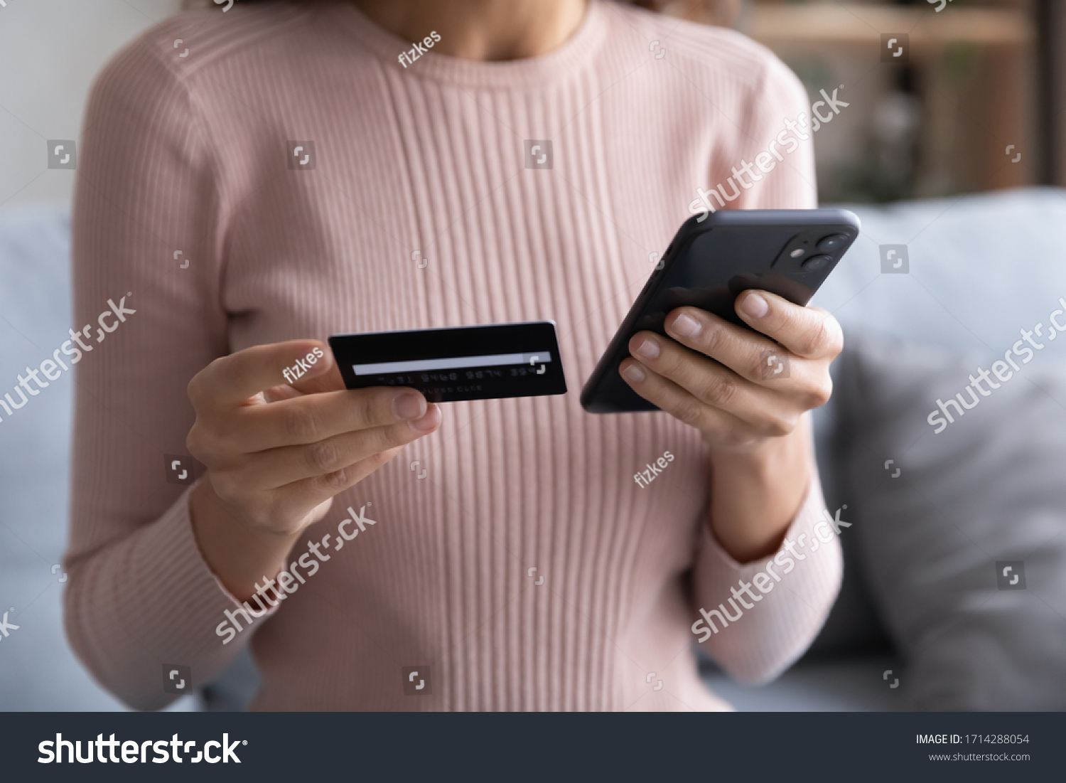 Close up young woman holding plastic credit card in one hand and telephone in another, entering cvv code, confirming purchase in online store. Female client using e-banking application on smartphone. #1714288054