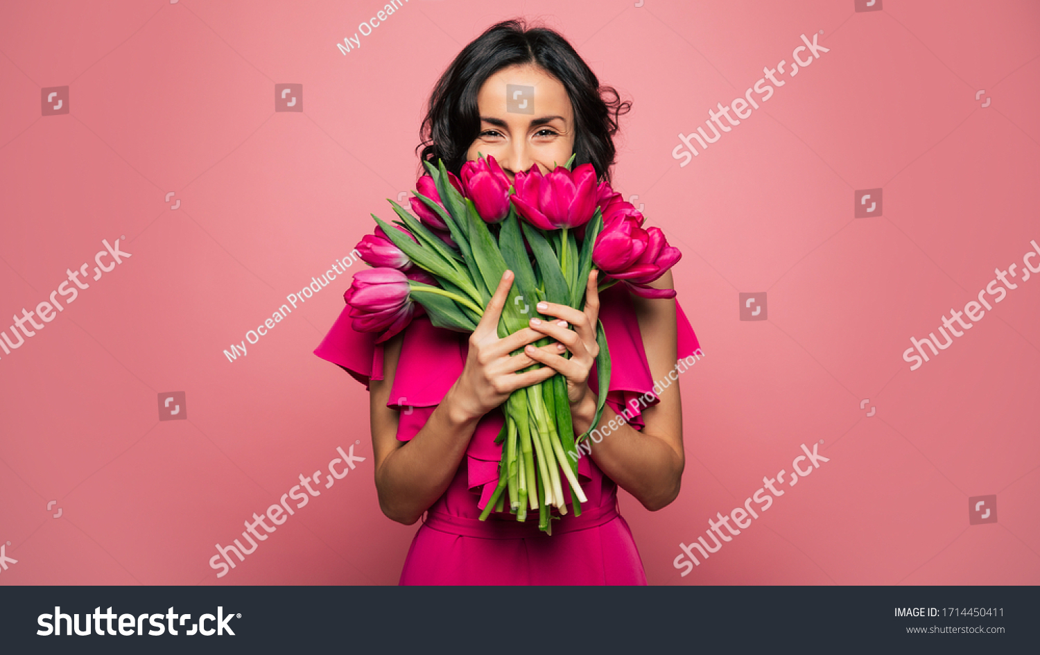 International Women's Day. Extremely happy woman in a bright pink dress is smelling a bunch of spring flowers, which she is holding in her hands. #1714450411