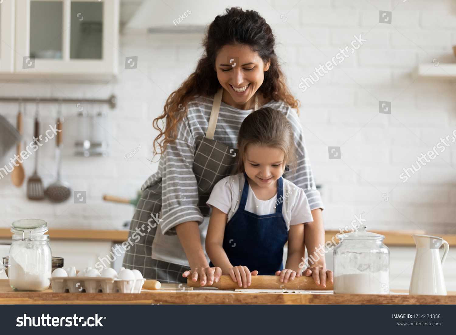 Happy attractive mommy helping cute smiling little preschool child daughter rolling dough for homemade pastry. Excited two female generations family enjoying cooking process together in kitchen. #1714474858