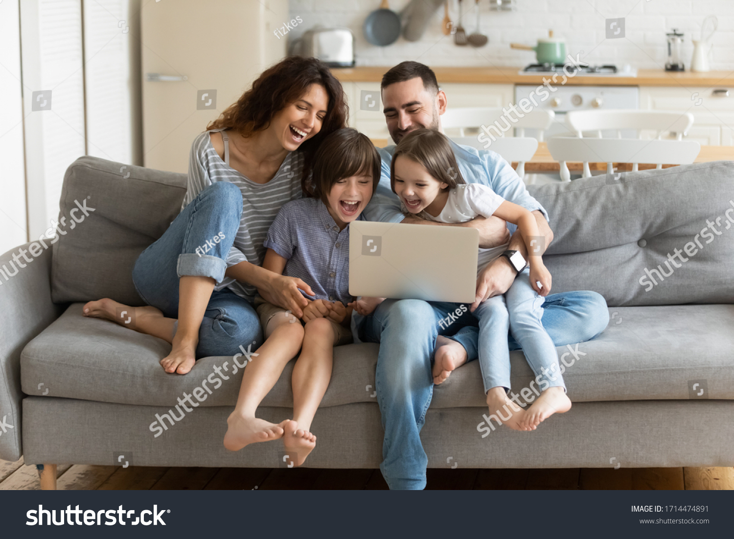 Overjoyed kids sitting on sofa with cheerful parents, watching funny video on computer. Happy married couple enjoying spending weekend time with small children, laughing looking at laptop screen. #1714474891