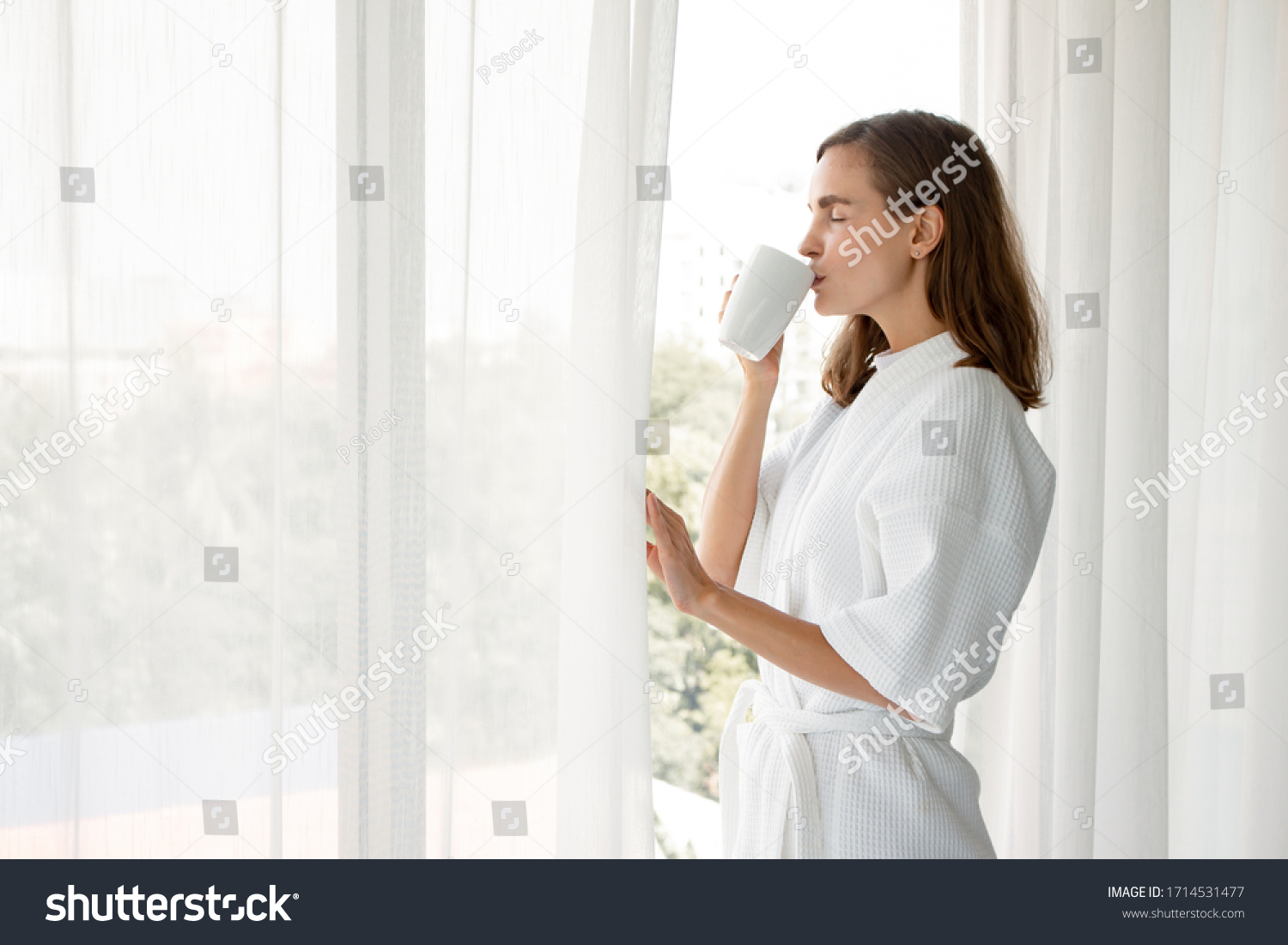 Beautiful Young Caucasian woman in pajamas holding a glass of white coffee ready to drink by the window in the bedroom at home after wake up in the morning. Woman standing drinking coffee by window. #1714531477