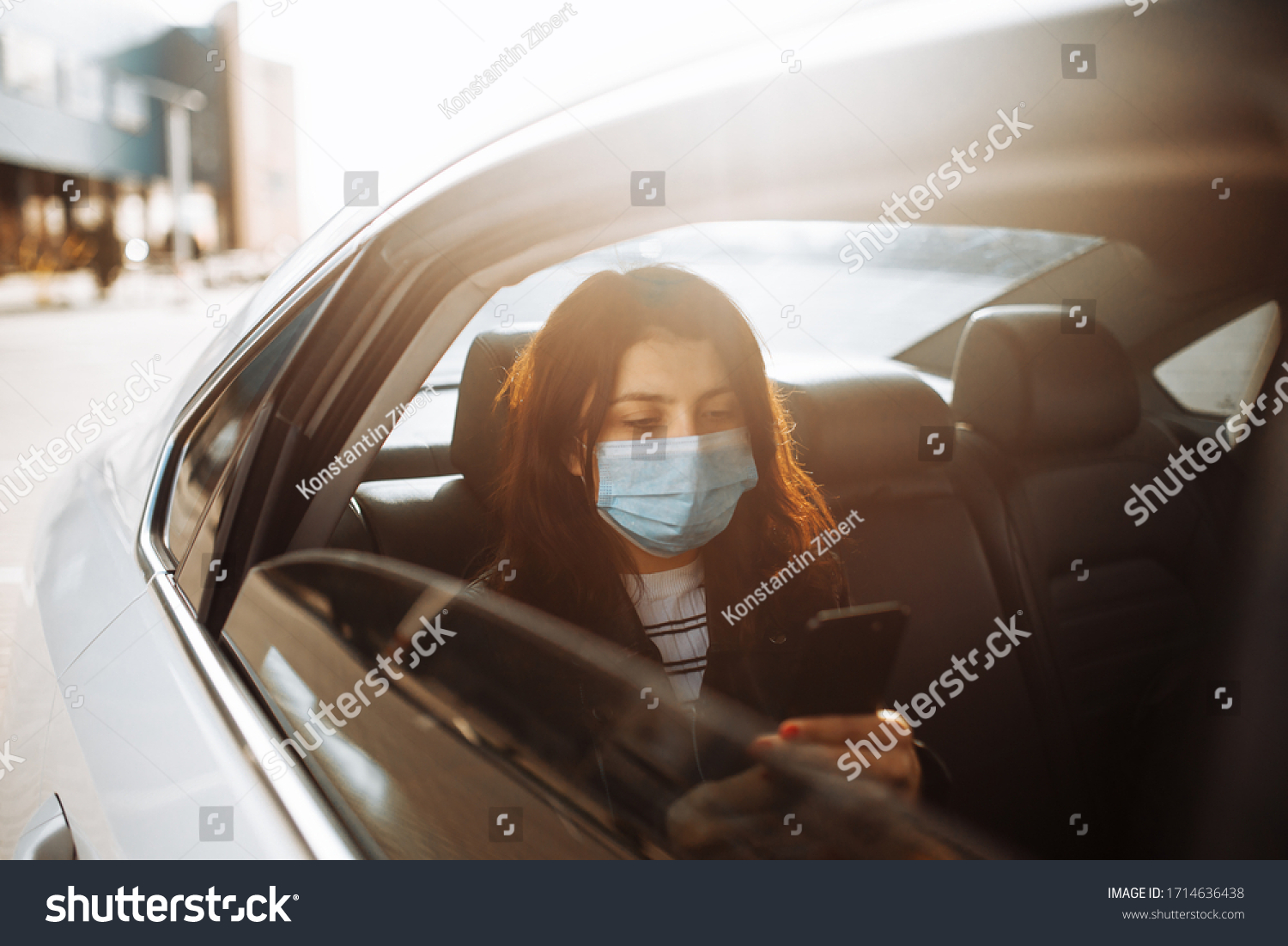 Woman wearing a medical sterile mask in taxi car on a backseat looking out of window checking her cell phone. Girl passenger waiting in a traffic jam during coronavirus quarantine. Healthcare concept #1714636438