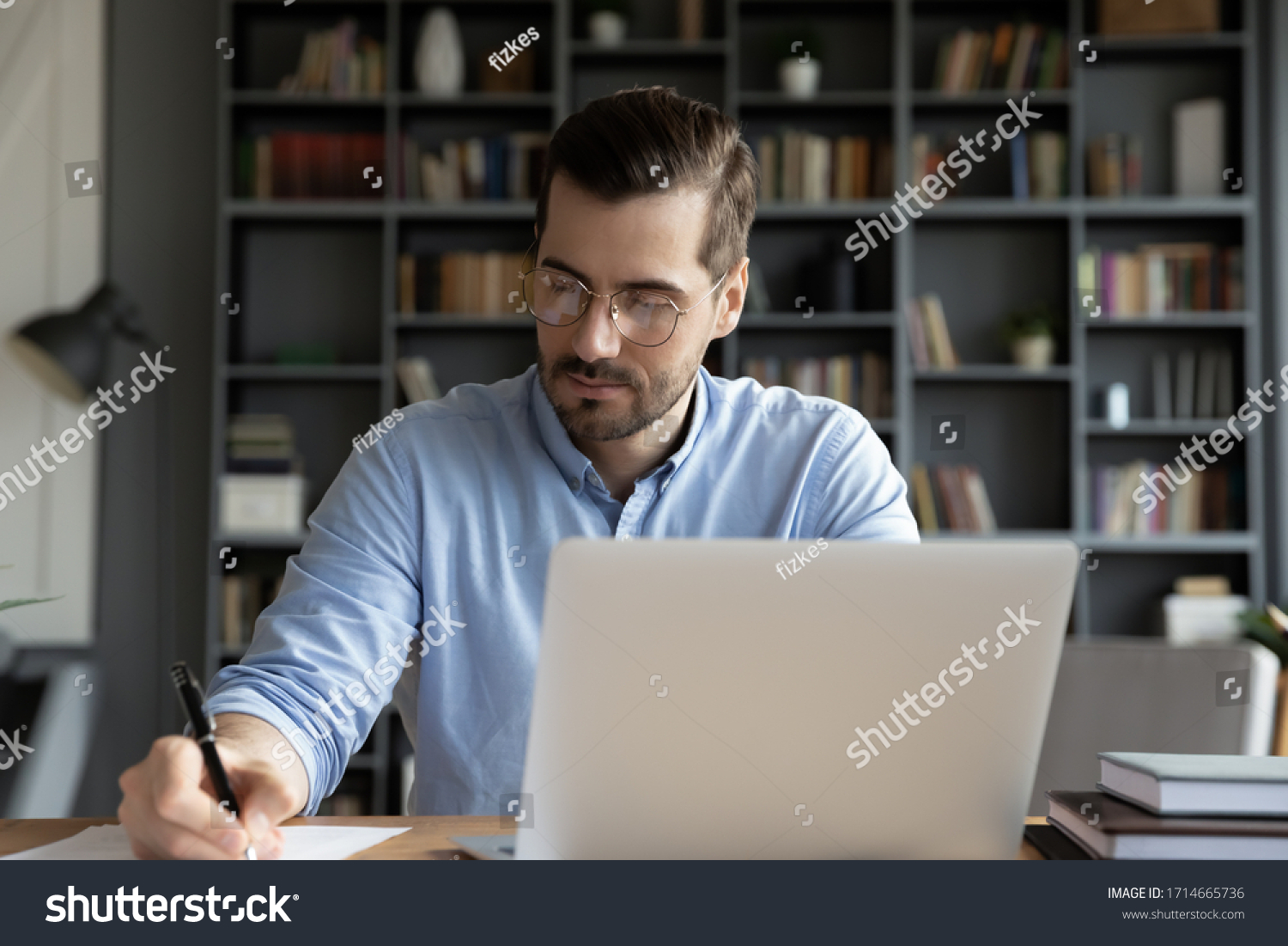 Confident businessman wearing glasses writing notes or financial report, sitting at desk with laptop, focused serious man working with paper documents, student studying online, research work #1714665736