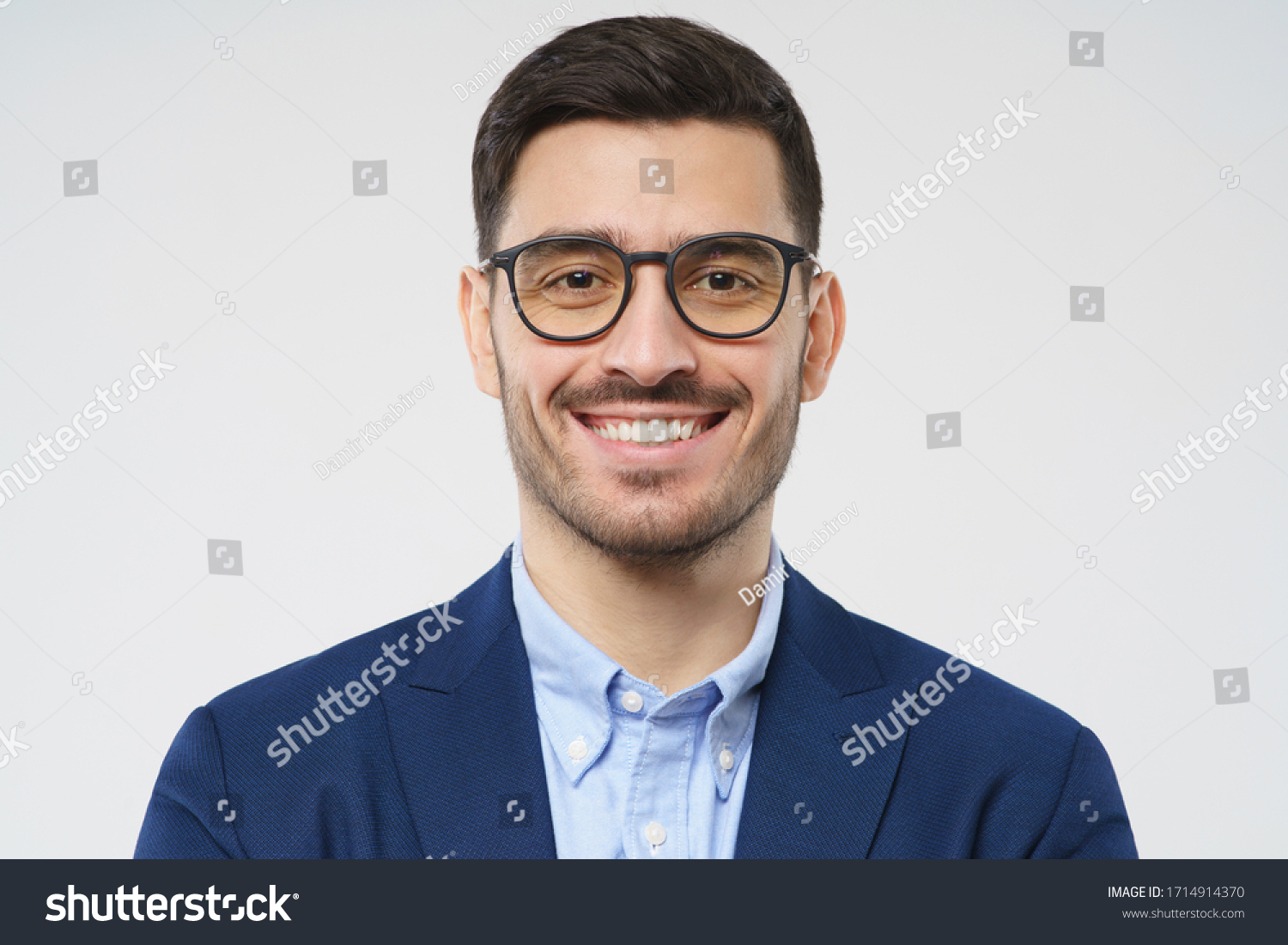 Close-up headshot of young businessman wearing glasses and smart casual suit, smiling at camera, isolated on gray background #1714914370