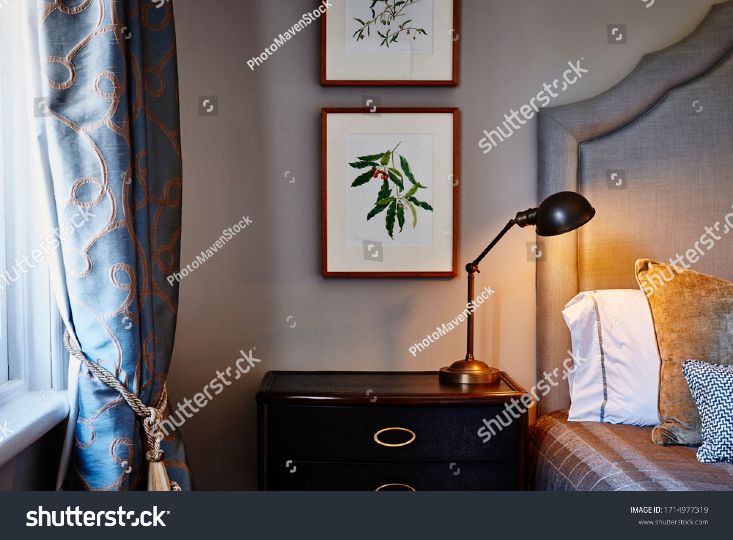 Interior photography of a loft bedroom in a Victorian mansion, a bed with an upholstered headboard, cushions, lamp, drapes and framed botanical illustrations #1714977319
