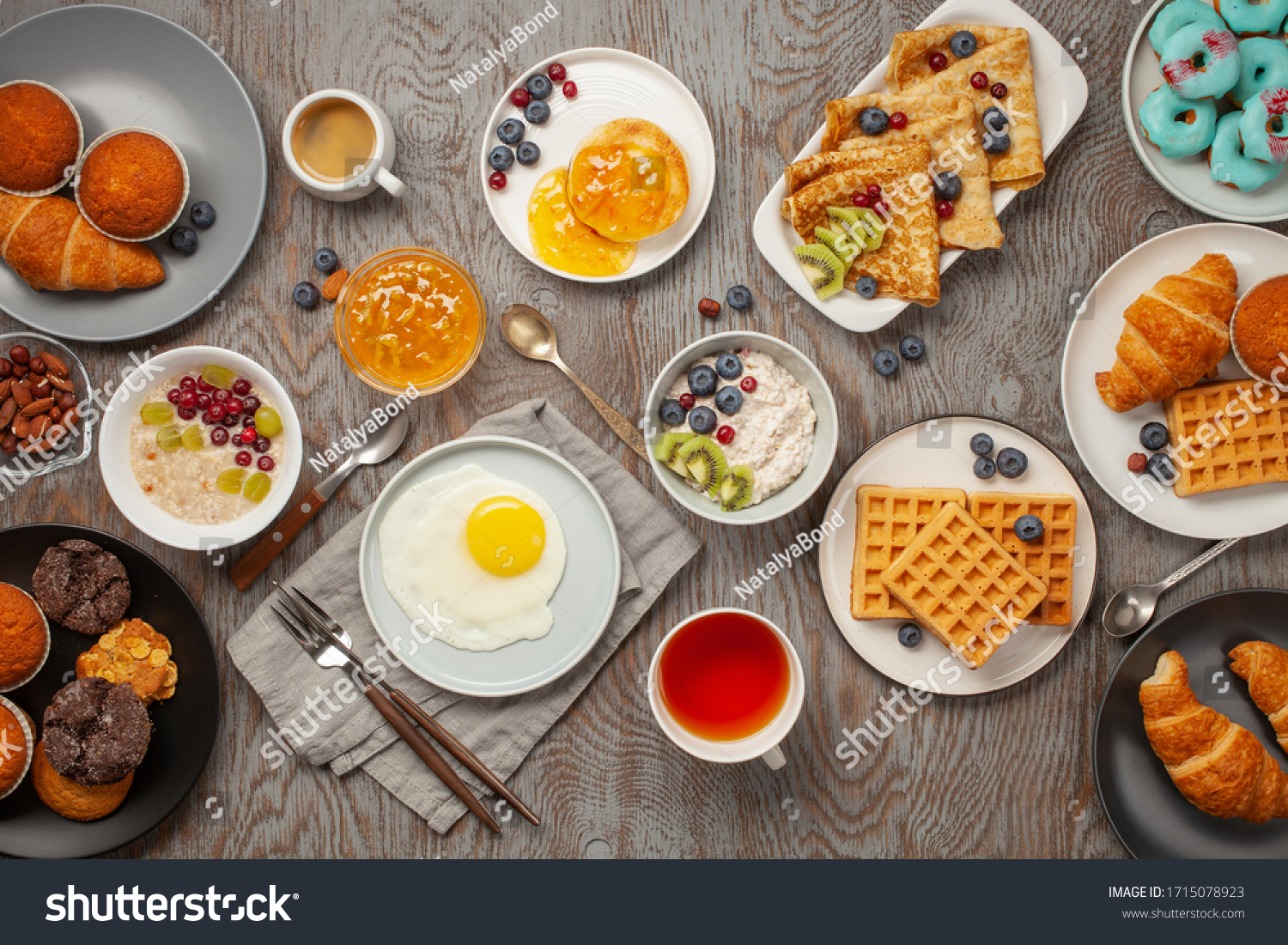 Continental breakfast captured from above (top view, flat lay). Coffee, tea, croissants, jam, egg, pancakes, maffins and oatmeal. Wooden background. Family breakfast table. #1715078923