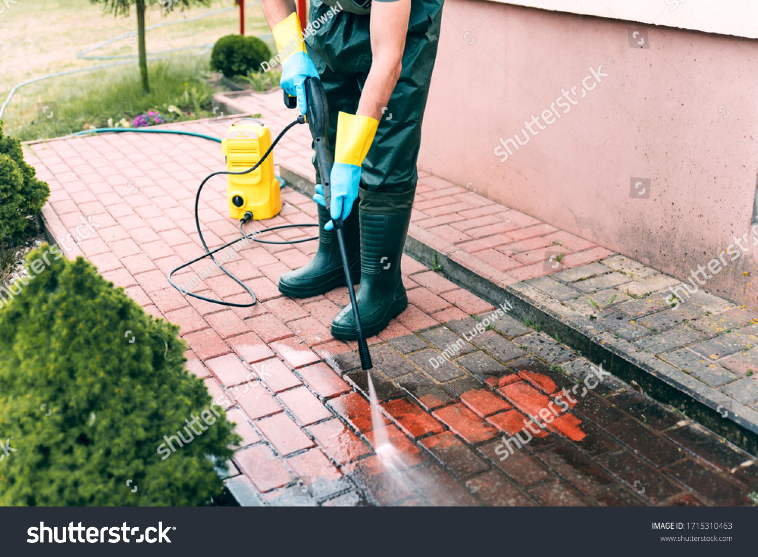 Man cleaning red concrete pavement blocks using high pressure water cleaner. Paving cleaning concept. Man wearing waders, protective waterproof trousers, doing spring jobs in garden. #1715310463
