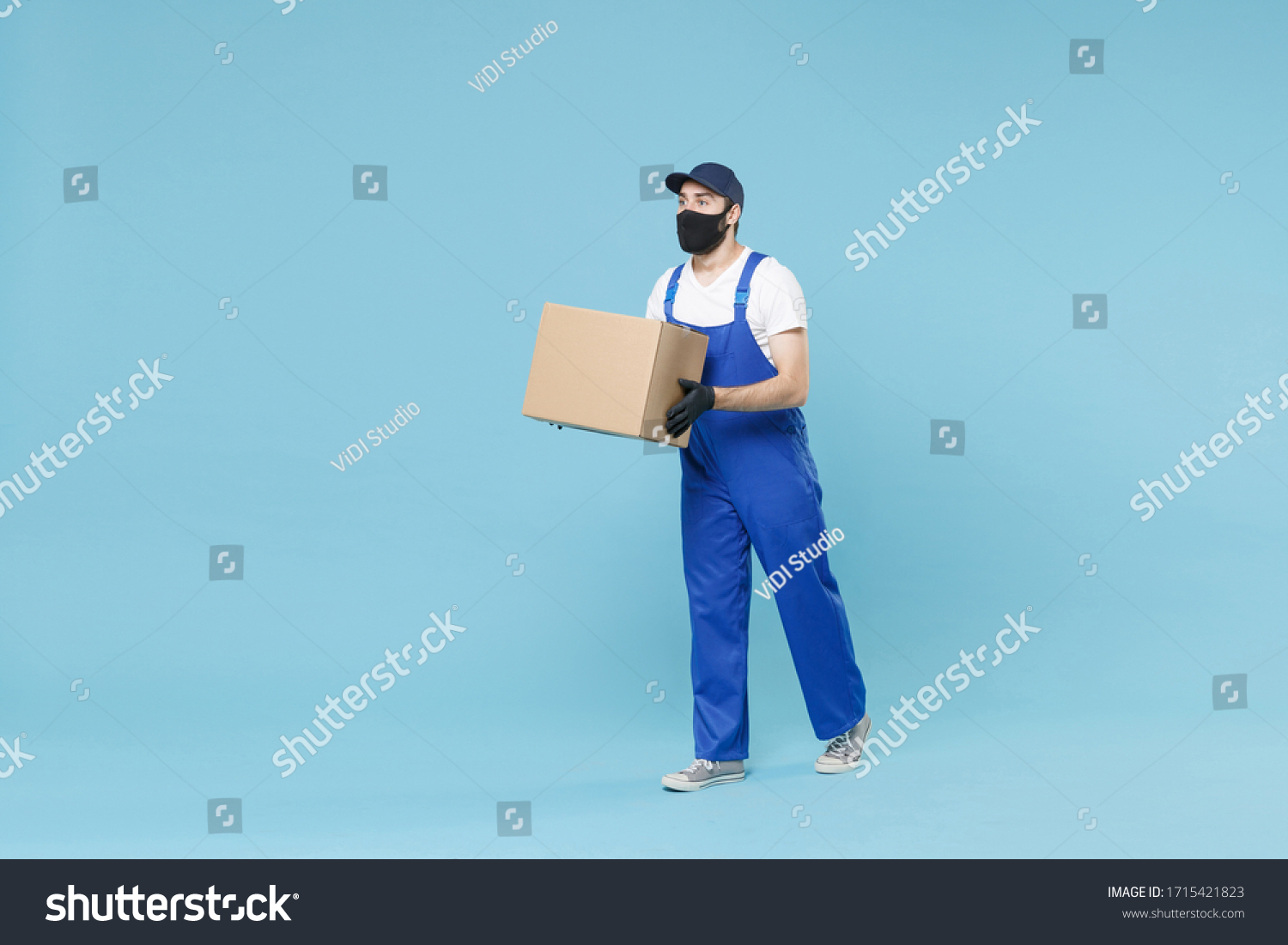 Full length delivery man in cap t-shirt uniform sterile face mask gloves isolated on blue background studio Guy employee courier hold box Service pandemic coronavirus virus covid-19 2019-ncov concept #1715421823