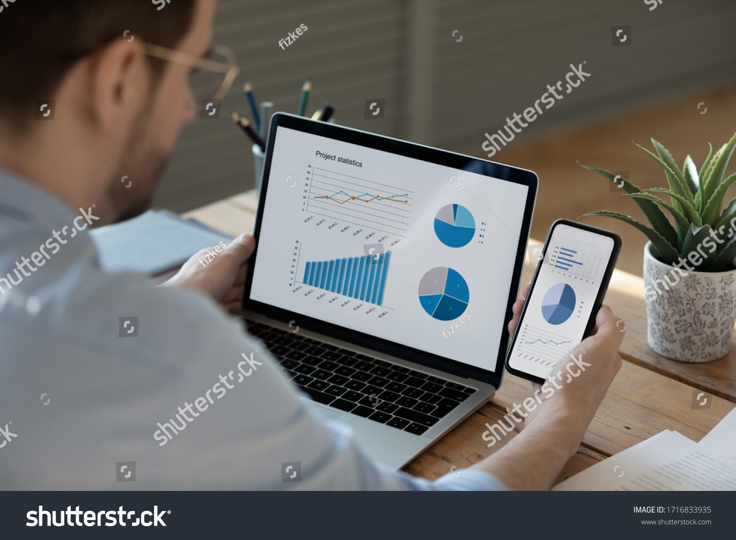 Close up businessman working with statistics, looking at laptop and phone screens with graphs and diagrams, synchronizing electronic devices, checking financial report presentation, sitting at desk #1716833935