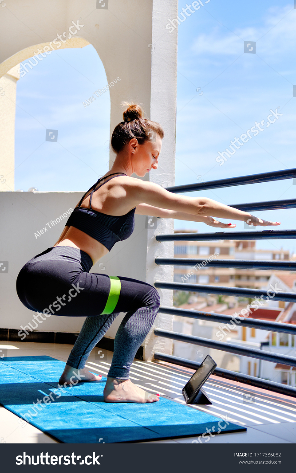 Work out at home due self-isolation COVID-19 corona virus pandemic concept. Woman in sportswear standing barefoot on mat perform squats with elastic band use tablet device video online lesson concept #1717386082