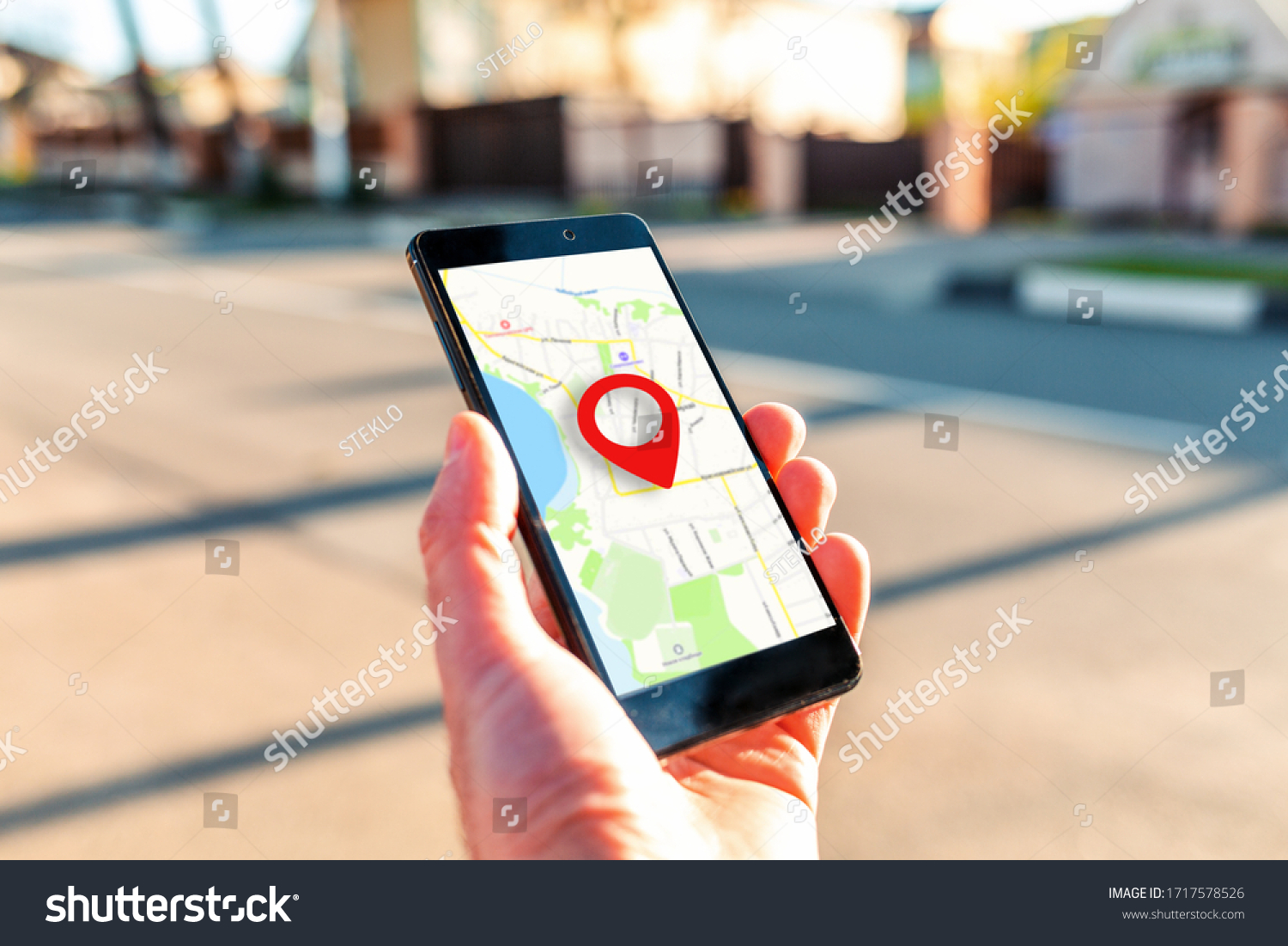 Male hand holding holding smartphone with online-a map on which the geolocation icon. In the background, a blurred street. Close up. Concept of online navigation and GPS #1717578526
