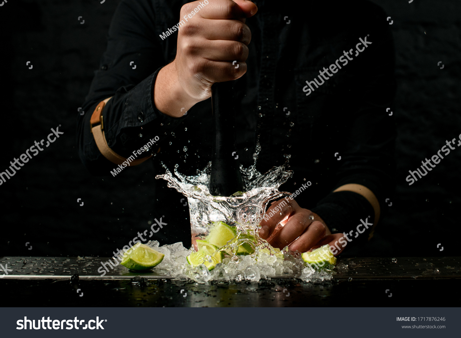 Close-up. Man bartender holds black madler in his hand and energetically squeeze slices of citrus to cocktail. Pieces of ice and lime at the bar. #1717876246