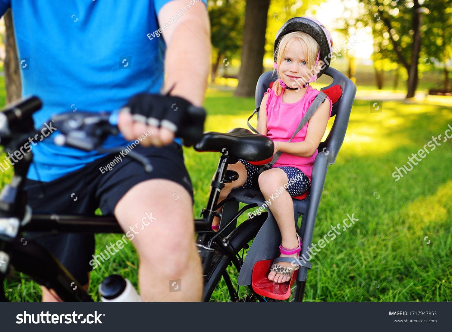 child a little girl in a Bicycle helmet smiles sitting in a child's Bicycle seat against the background of a Park and green grass. #1717947853