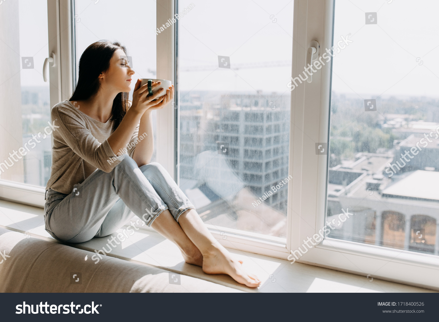 Young woman looking through the window with a city view, sitting on a windowsill, enjoying coffee or tea in the morning. #1718400526