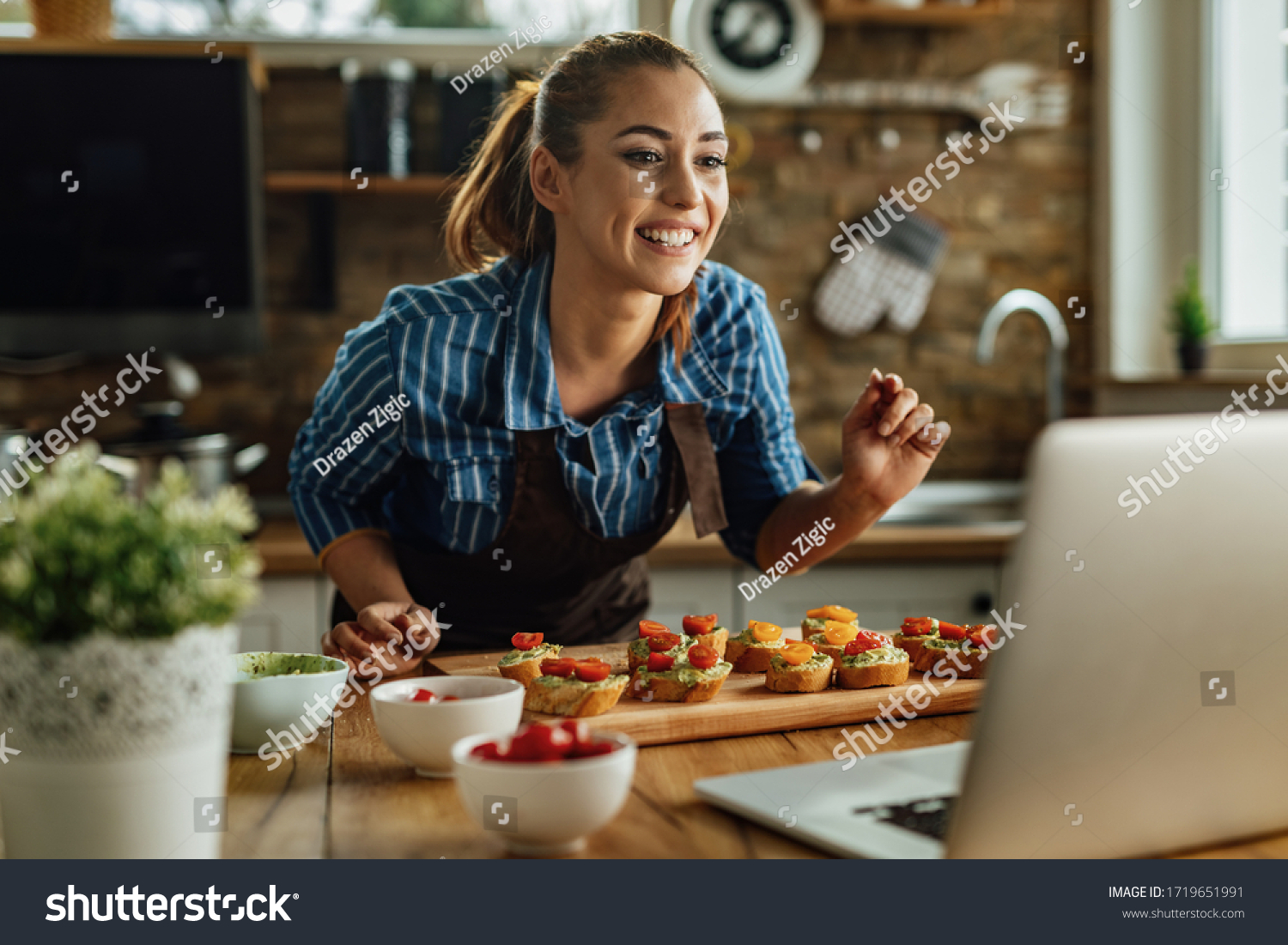 Young smiling woman preparing food in the kitchen while live streaming over laptop in the kitchen.  #1719651991