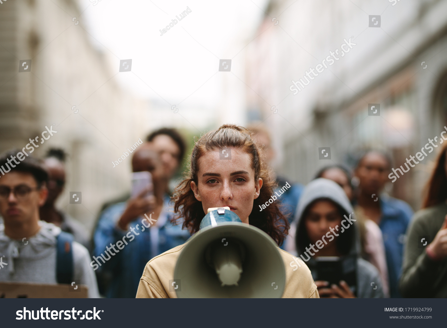 Young woman with a megaphone with group of demonstrator in background. Woman protesting with megaphone in the city. #1719924799
