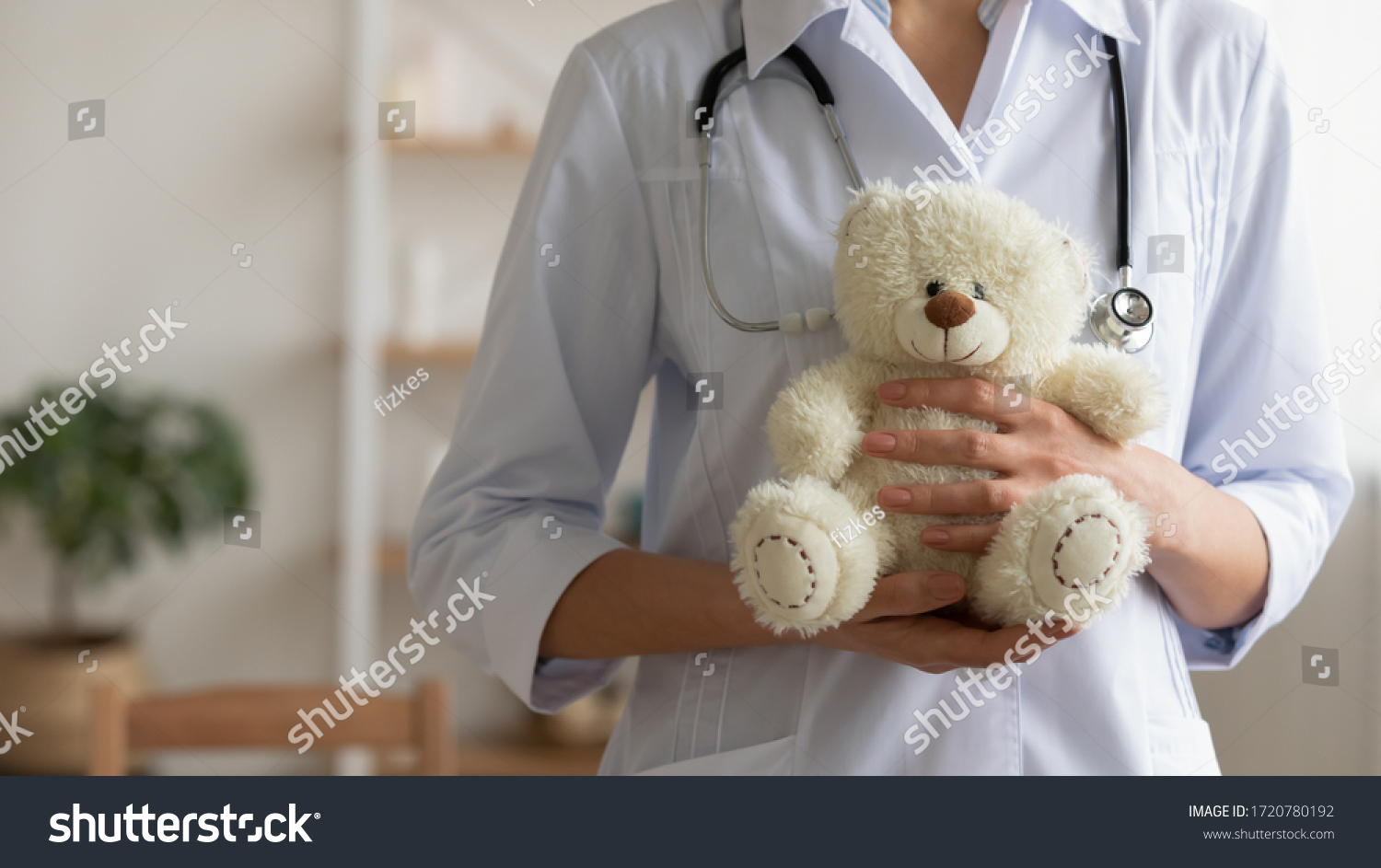 Female pediatrician holding teddy bear in hands. Professional paediatrician doctor wears white coat standing with toy. Children healthcare paediatric medical services background concept. Close up view #1720780192