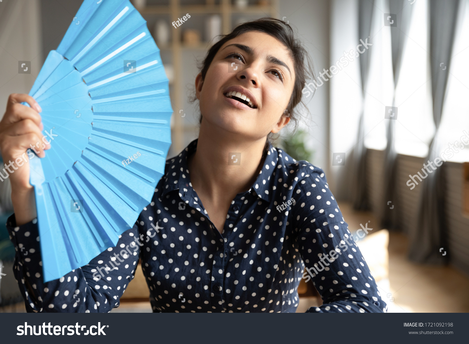 Head shot young indian woman using blue paper fan, feeling hot relieved indoors. Sweaty millennial hindu girl teenage cooling herself, breathing fresh air, spending time at home without conditioner. #1721092198