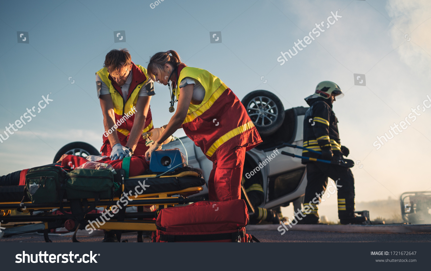 On the Car Crash Traffic Accident Scene: Paramedics Saving Life of a Female Victim who is Lying on Stretchers. They Apply Oxygen Mask, Do Cardiopulmonary Resuscitation / CPR and Perform First Aid #1721672647