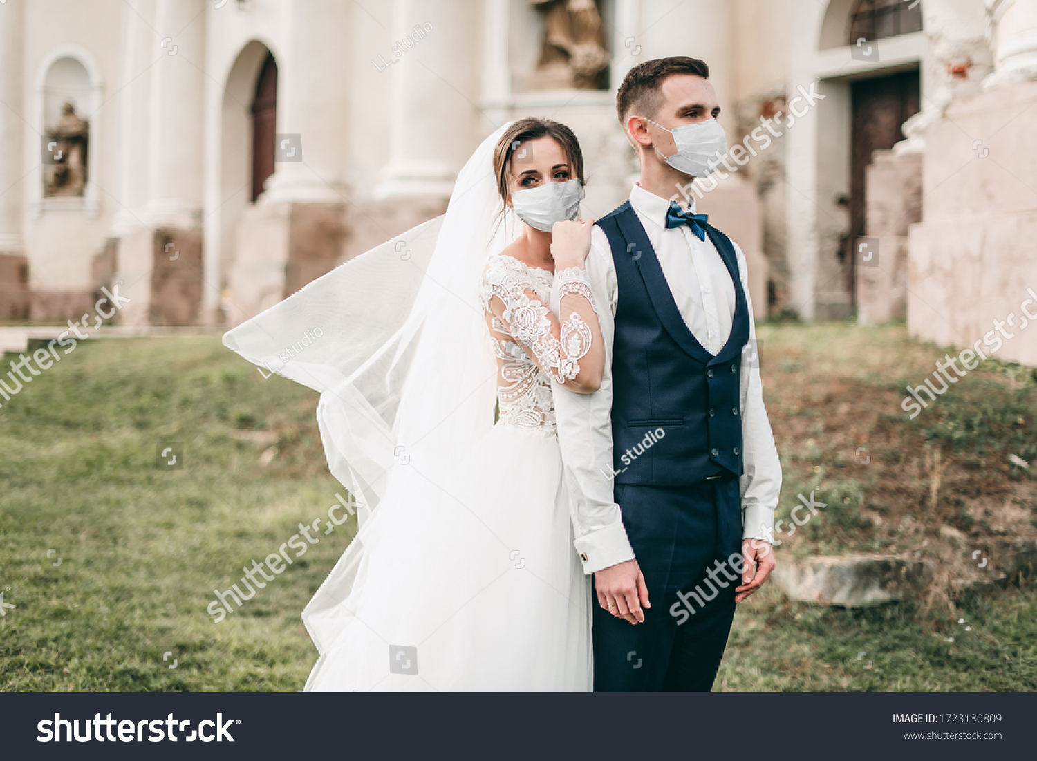 Young loving brides walking and hugging in the city near the church in medical masks with a bouquet of roses during quarantine on their wedding day. Coronavirus, disease, protection, illness, corona #1723130809