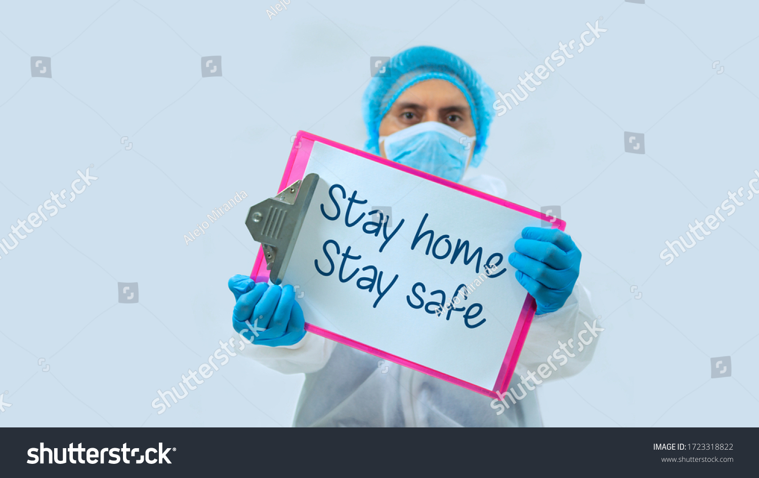 Doctor with face mask and blue gloves in background, holding a clipboard with white sheets with the message: STAY HOME, STAY SAFE in the foreground #1723318822