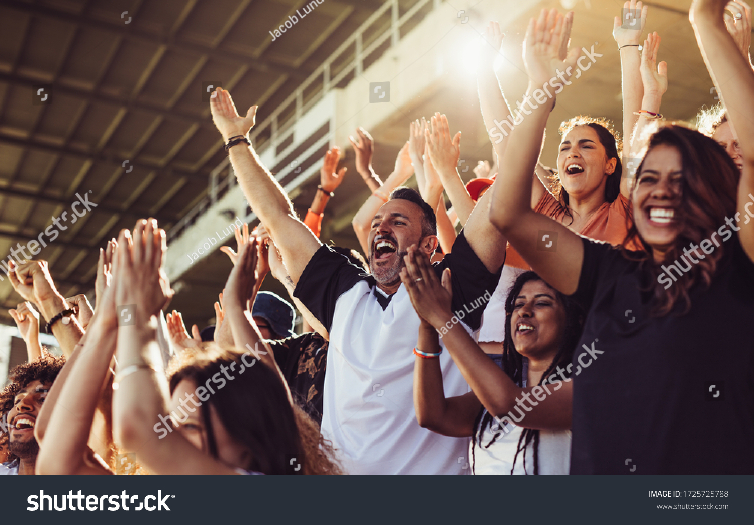 Crowd of sports fans cheering during a match in stadium. Excited people standing with their arms raised, clapping and yelling to encourage their team. #1725725788