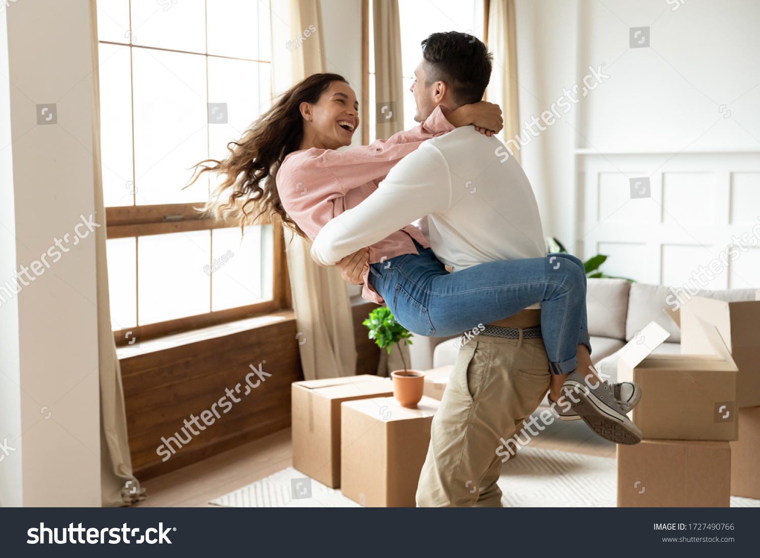Loving husband lifting excited wife, celebrating moving day, having fun in modern living room with cardboard boxes with belongings, happy young couple purchasing new house, mortgage and relocation #1727490766