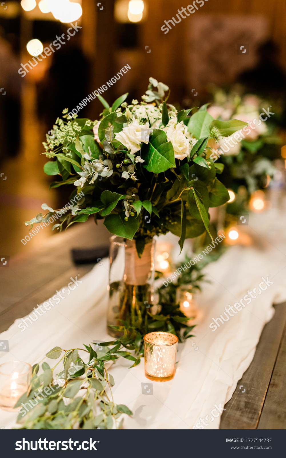 Bridal Bouquet with White Roses, Baby's Breath and Eucalyptus Leaves in a Vase at the Head Table at a Wedding with Votive Candles and a White Cheesecloth on a Wood Table #1727544733