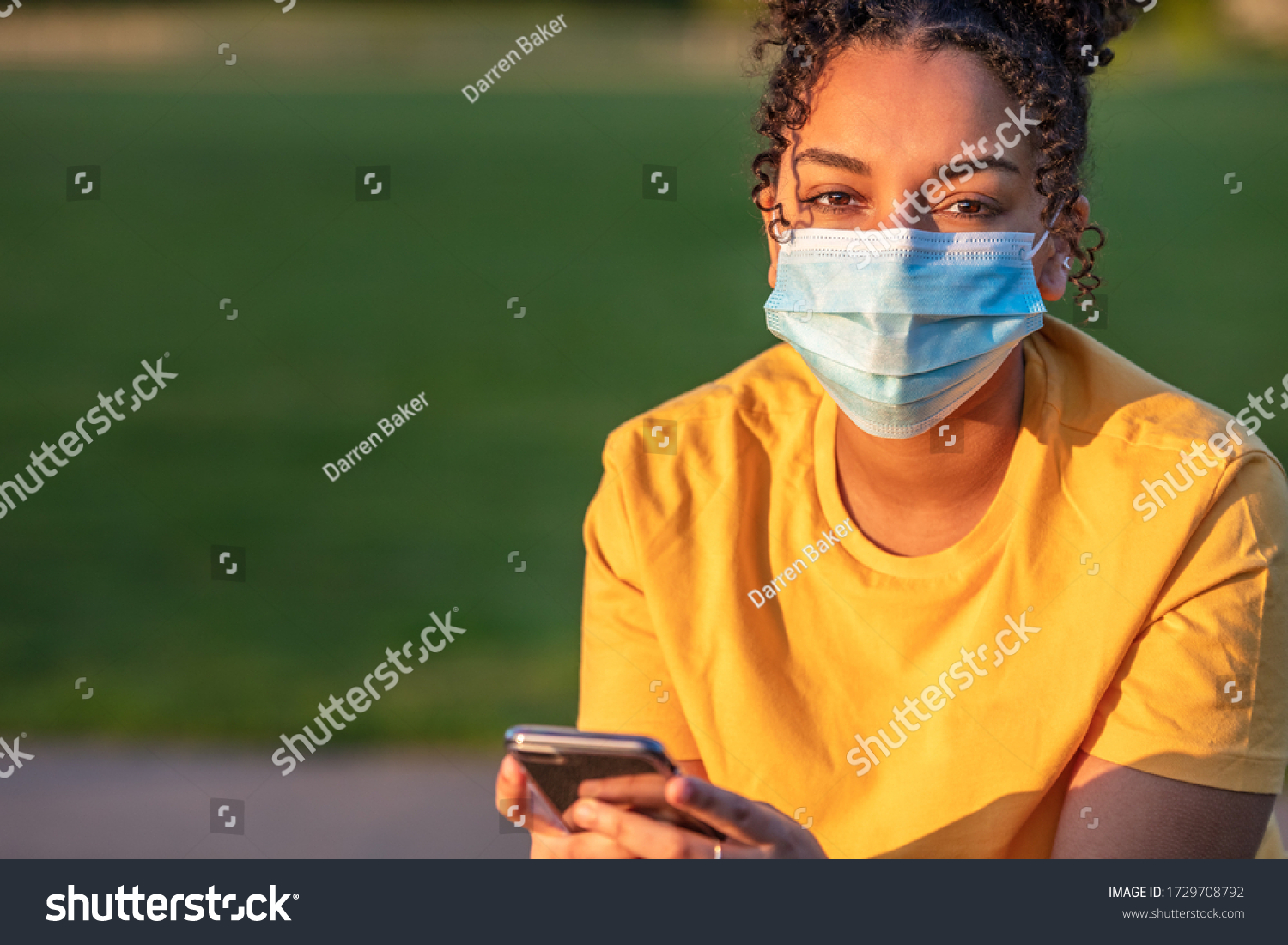 Mixed race biracial African American teenager teen girl young woman wearing a face mask and using mobile cell phone outside during the Coronavirus COVID-19 virus pandemic #1729708792