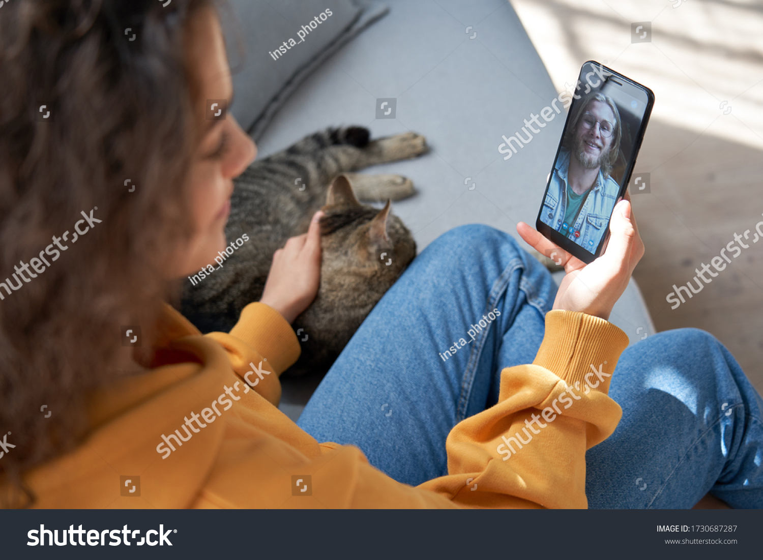 Young hispanic latin teen girl relax sit on sofa with cat at home holding phone video calling distance friend dating online on mobile screen using smartphone videochat app. Over shoulder closeup view #1730687287
