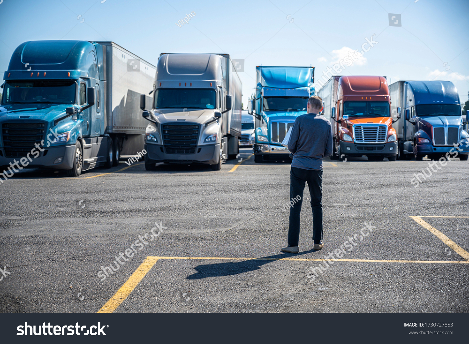 Slim Truck driver carries a box with purchases in his hands and going to his big rig semi ruck parked on the truck stop parking lot standing in row with another semi trucks with semi trailers #1730727853