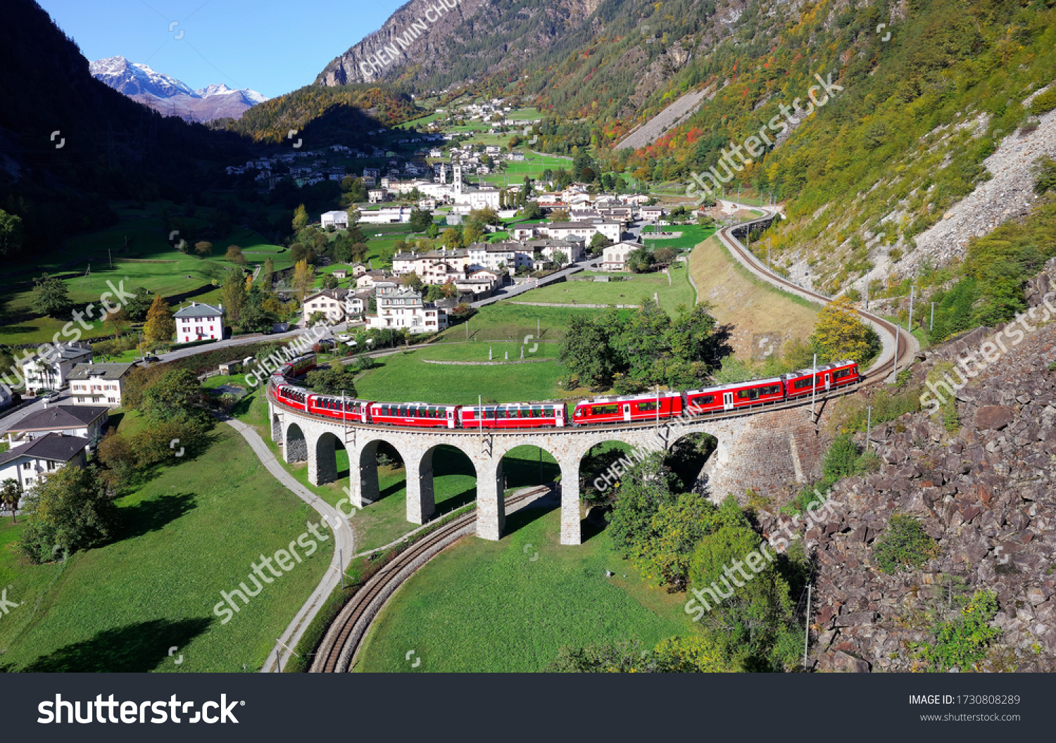 Aerial view of a Bernina Express train crossing the famous Brusio spiral viaduct of Rhaetian Railway over a green grassy meadow with fall colors on the rocky mountainside, in Graubunden, Switzerland #1730808289