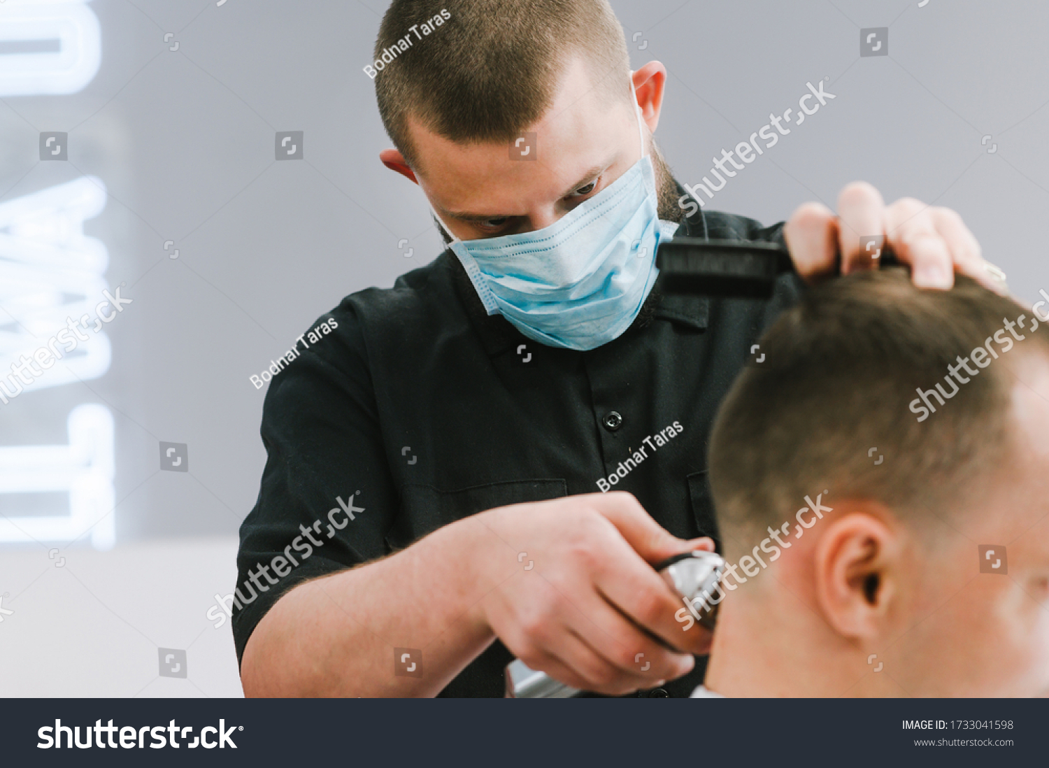 Professional barber in a medical gauze mask on the face cuts the client's hair with a clipper with a focused gaze. Hairdresser works in quarantine. Close up #1733041598