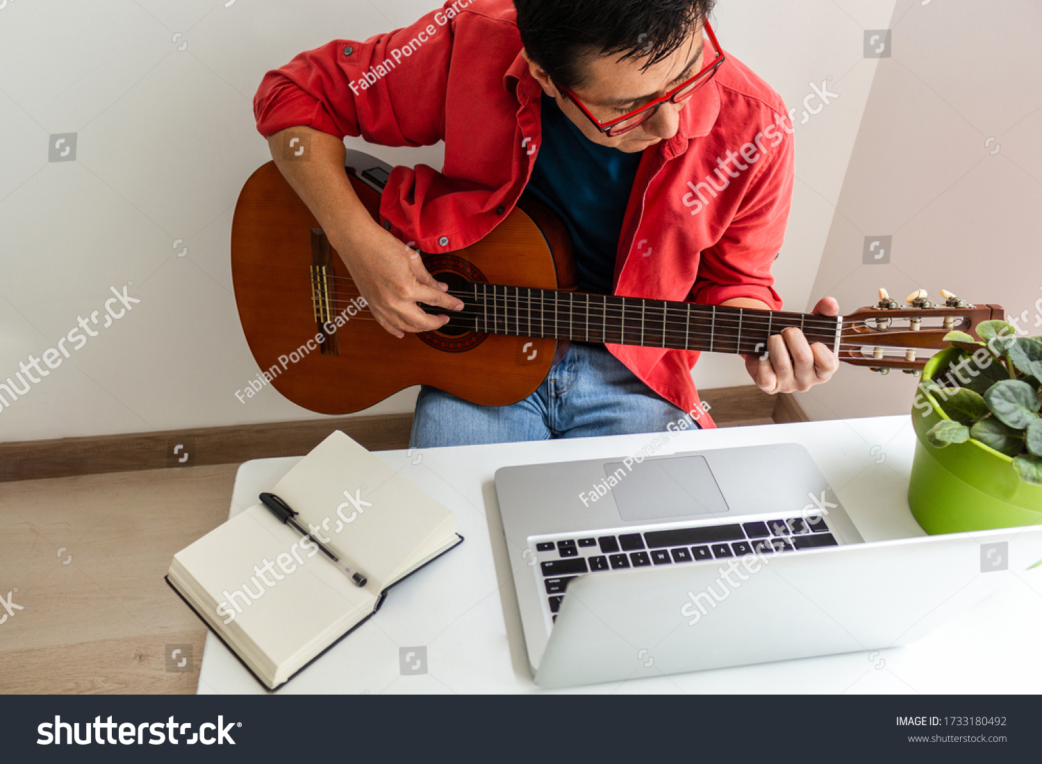In a room at home a man plays the guitar in front of his laptop during an online class #1733180492