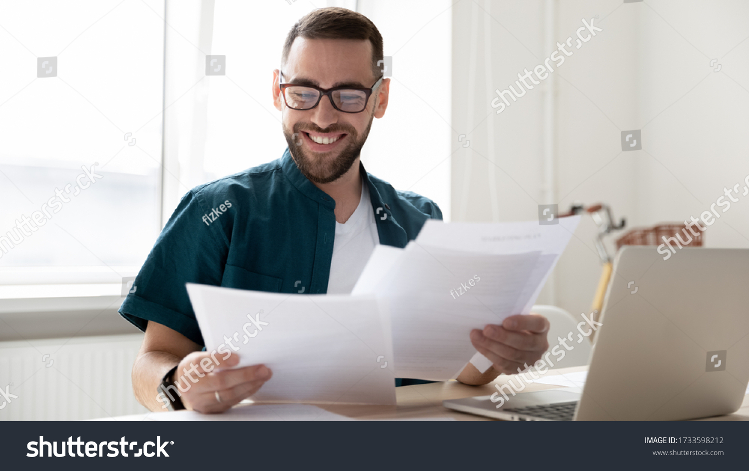 Happy handsome businessman holding and reading documents with good news near laptop. Young smiling bearded man, student rejoice and celebrate successful distance deal for work. #1733598212