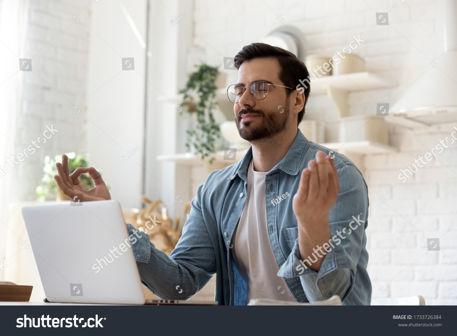 Calm millennial Caucasian man in glasses sit at table distracted from computer work meditate in kitchen, peaceful young male practice yoga relieve negative emotions at workplace, stress free concept #1733726384