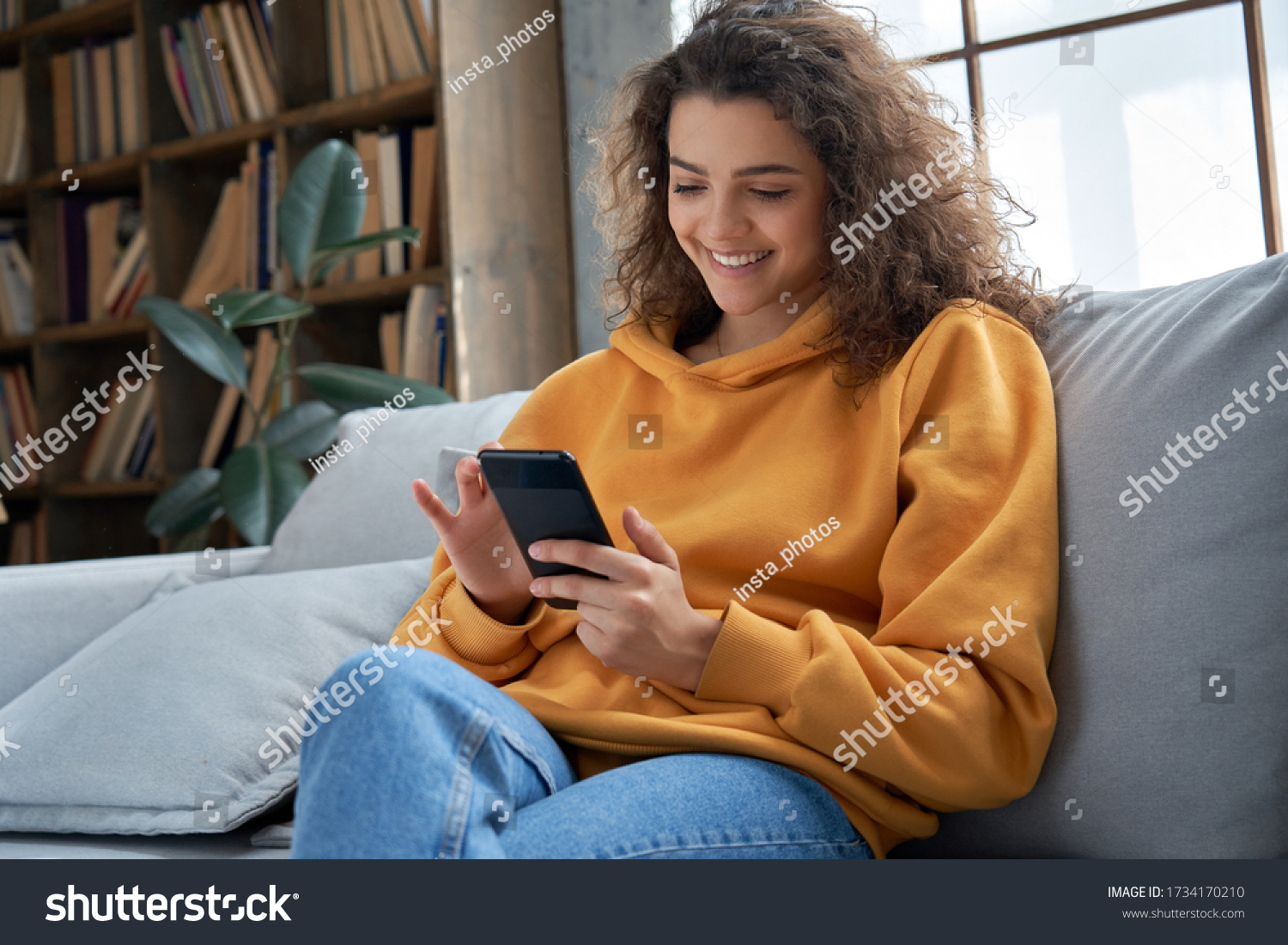 Happy millennial hispanic teen girl checking social media holding smartphone at home. Smiling young latin woman using mobile phone app playing game, shopping online, ordering delivery relax on sofa. #1734170210