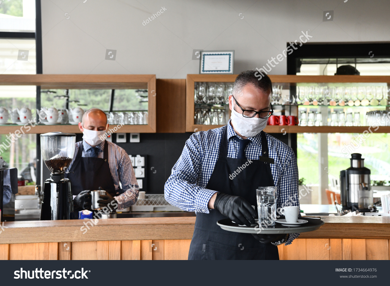 waiter in a medical protective mask serves  the coffee in restaurant durin coronavirus pandemic representing new normal concept #1734664976