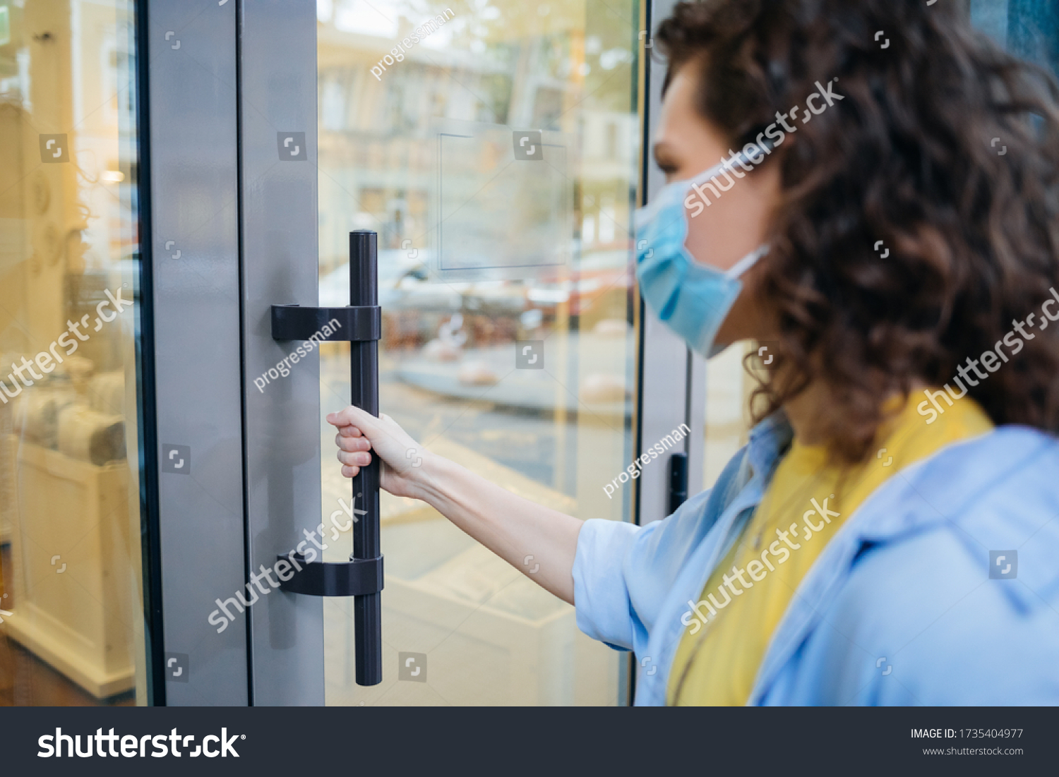 Young woman wearing protective medical mask holds on to the handle and opens the door when entering store. #1735404977