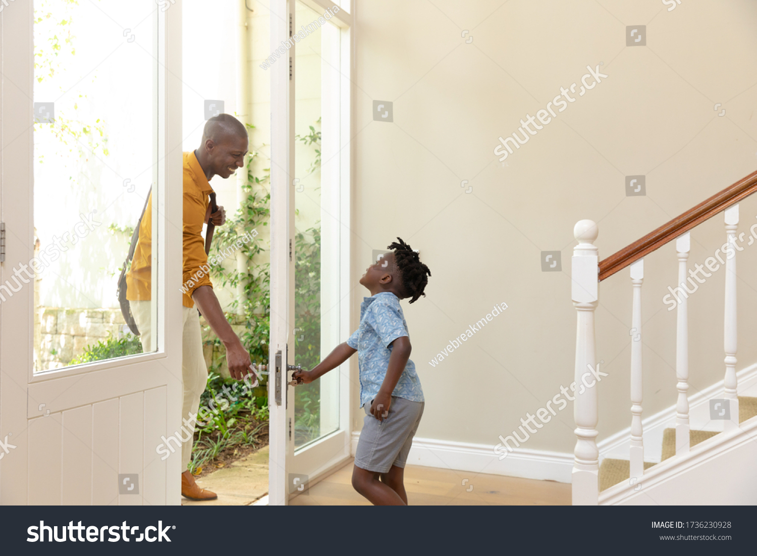 Side view of an African American man arriving home, welcomed by his young son opening the front door. Social distancing and self isolation in quarantine lockdown for Coronavirus Covid19 #1736230928
